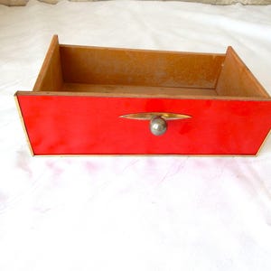 Può includere: Un cassetto rettangolare in legno caratterizzato da un pannello frontale rosso vivace con un bordo metallico color oro. Il cassetto include un pomello sferico color argento centrato, montato su una piastra metallica orizzontale a mandorla.