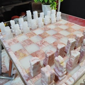 Pu&ograve; includere: Un set di scacchi in marmo bianco e rosa con pezzi bianchi su una scacchiera rosa. I pezzi sono scolpiti in uno stile unico, che ricorda piccole torri.