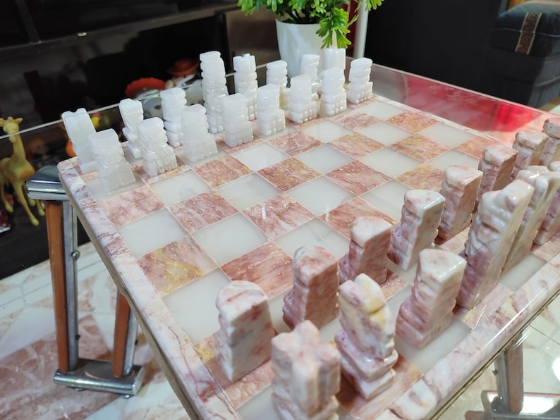 Pu&ograve; includere: Un set di scacchi in marmo bianco e rosa con pezzi bianchi su una scacchiera rosa. I pezzi sono scolpiti in uno stile unico, che ricorda piccole torri.