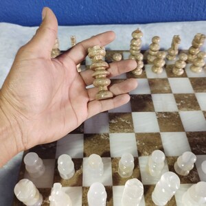 May include: A hand holds a white chess piece, a queen, on a marble chessboard with brown and white squares. The board is set up with other white chess pieces.