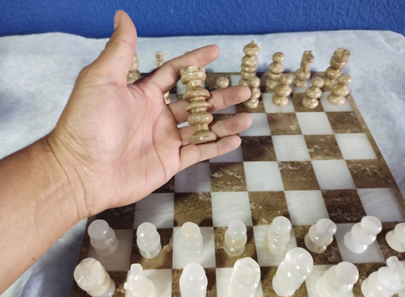 May include: A hand holds a white chess piece, a queen, on a marble chessboard with brown and white squares. The board is set up with other white chess pieces.