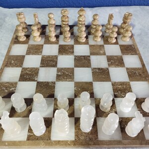 May include: A white marble chess set with brown marble accents. The chess pieces are arranged on a brown and white marble chessboard. The chessboard is made of a light brown marble with white marble squares.