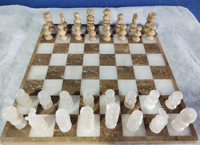 May include: A white marble chess set with brown marble accents. The chess pieces are arranged on a brown and white marble chessboard. The chessboard is made of a light brown marble with white marble squares.