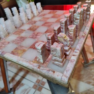 Pu&ograve; includere: Un set di scacchi in marmo con pezzi rosa e bianchi su una scacchiera in marmo rosa e bianco.