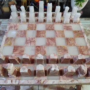 Pu&ograve; includere: Un set di scacchi in marmo bianco con una scacchiera in marmo rosa e bianco. I pezzi degli scacchi sono scolpiti in uno stile tradizionale e la scacchiera ha un motivo a scacchiera.