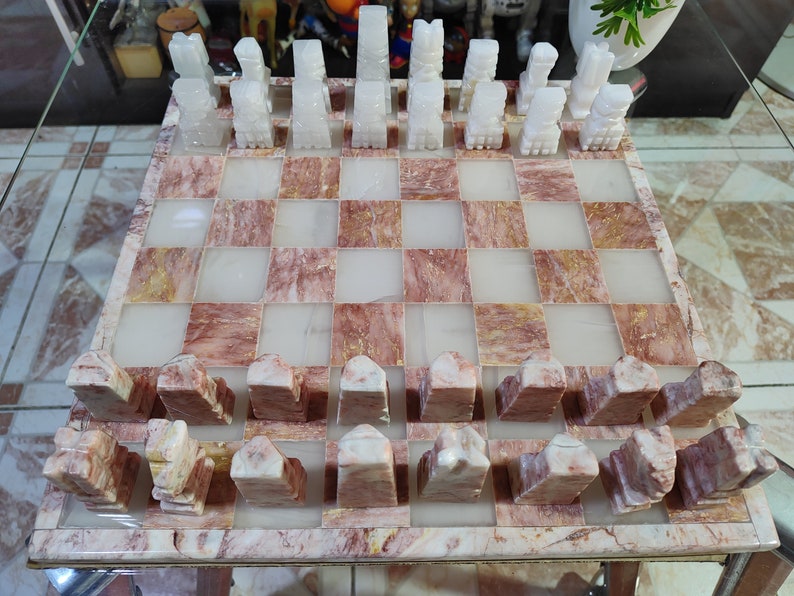 Pu&ograve; includere: Un set di scacchi in marmo bianco con una scacchiera in marmo rosa e bianco. I pezzi degli scacchi sono scolpiti in uno stile tradizionale e la scacchiera ha un motivo a scacchiera.