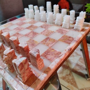 Pu&ograve; includere: Un set di scacchi in marmo rosa e bianco con pezzi bianchi sulla scacchiera e pezzi rosa fuori dal tabellone.