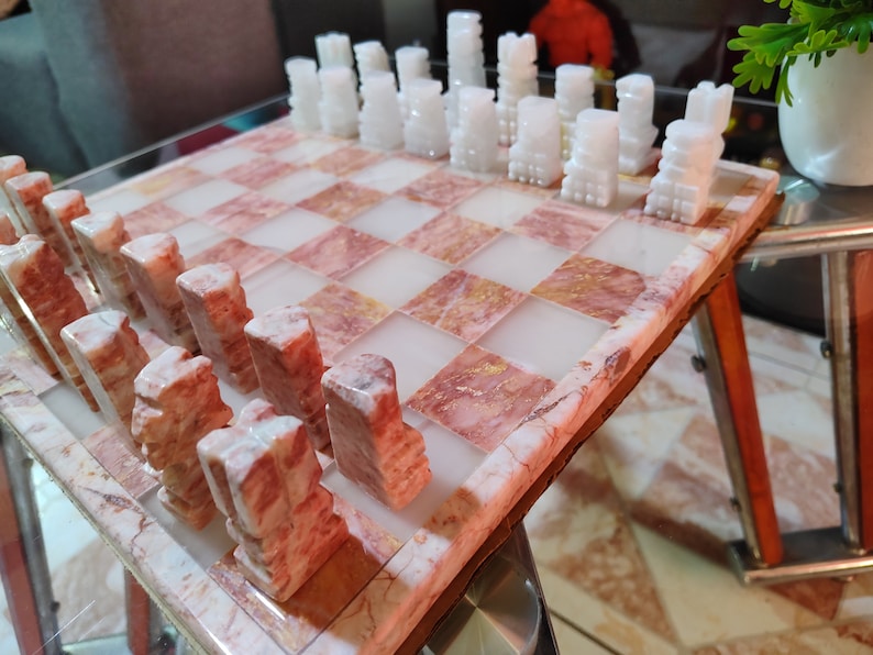 Pu&ograve; includere: Un set di scacchi in marmo rosa e bianco con pezzi bianchi sulla scacchiera e pezzi rosa fuori dal tabellone.