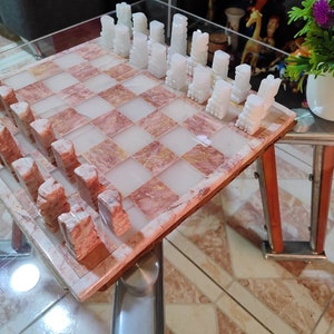Pu&ograve; includere: Un set di scacchi in marmo con pezzi bianchi e rosa su una scacchiera in marmo rosa e bianco. La scacchiera &egrave; su un tavolo di vetro con una cornice in legno.