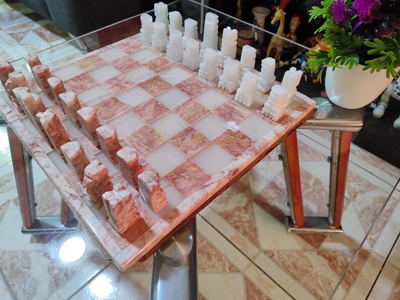 Pu&ograve; includere: Un set di scacchi in marmo con pezzi bianchi e rosa su una scacchiera in marmo rosa e bianco. La scacchiera &egrave; su un tavolo di vetro con una cornice in legno.