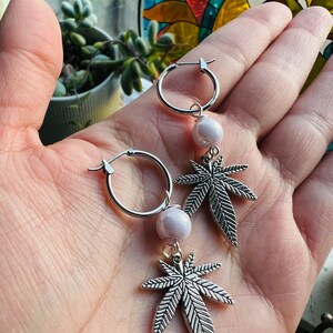 May include: A pair of silver hoop earrings with a dangling silver marijuana leaf charm and a white pearl bead.