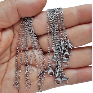 May include: Close-up of several silver chain necklaces with lobster claw clasps and jump rings. The chains are held in a hand, highlighting their delicate links and shiny finish. Suitable for jewellery making or crafting.