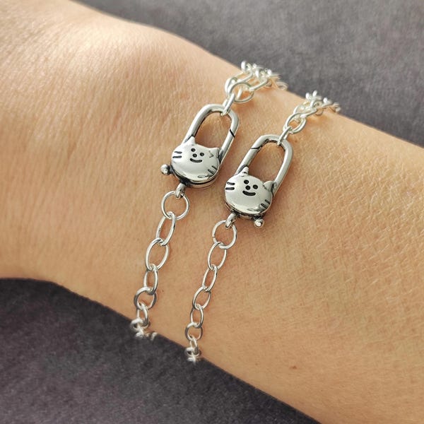 May include: Two silver-toned cat charm bracelets. Each bracelet features a cat-shaped clasp with a smiling face and a chain link design. The bracelets are worn on a person's wrist, showcasing the jewelry's design and detail.