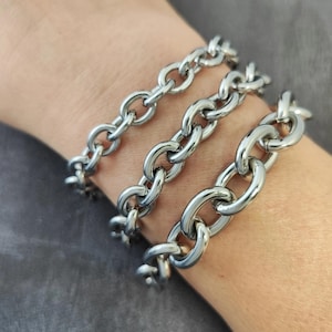 May include: Three silver chain bracelets with large oval links are worn on a wrist. The bracelets are made of shiny, polished metal and are arranged side by side. The background is a solid, neutral colour.