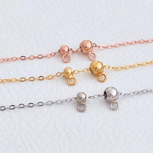 May include: Close-up of three delicate necklaces in rose gold, gold, and silver. Each necklace features a small ball charm and a chain with a clasp. The jewellery is displayed on a white surface.