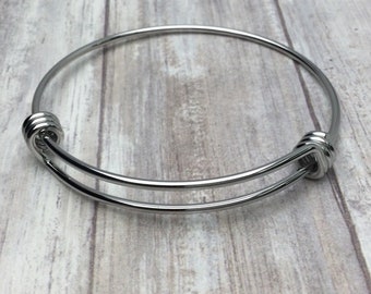May include: A silver-toned expandable wire bangle bracelet featuring a smooth, polished metallic finish. The circular design includes two overlapping ends that are coiled into small loops, allowing the bracelet to adjust in size.
