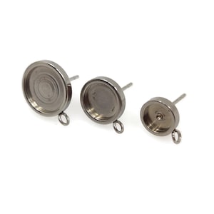 May include: Three silver-toned metal earring settings with a loop on the side. The settings are different sizes, with the largest setting being on the left and the smallest setting on the right.