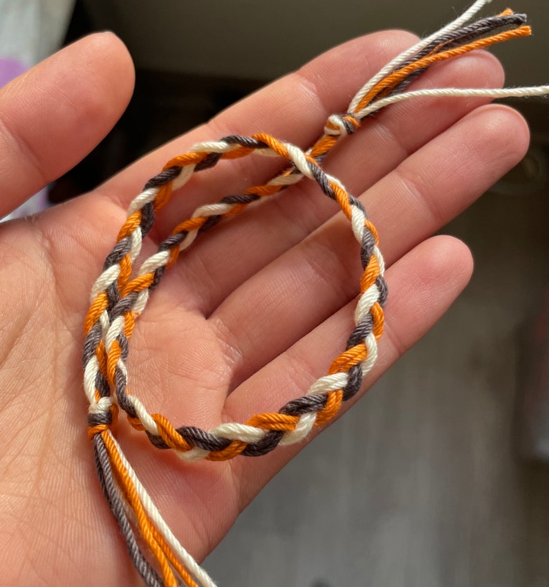 K&ouml;nnte beinhalten: Ein geflochtenes Freundschaftsarmband mit orangefarbenen, wei&szlig;en und braunen F&auml;den.