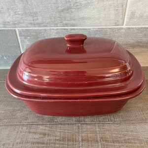 Puede incluir: Un plato para hornear de cerámica rojo con tapa. El plato tiene una forma rectangular con esquinas redondeadas y un borde ligeramente elevado. La tapa tiene un pequeño pomo en la parte superior.