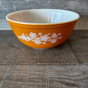 May include: An orange mixing bowl with a white floral design. The bowl has a white interior and a rounded shape. The floral pattern is a stylized cluster of flowers and leaves. The bowl is likely made of ceramic or glass.