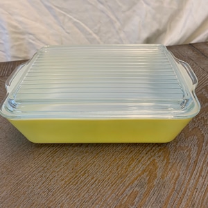 May include: A square, yellow Pyrex baking dish with a clear glass lid. The lid has a ribbed design. The dish is sitting on a wooden surface. The dish has handles on either side.
