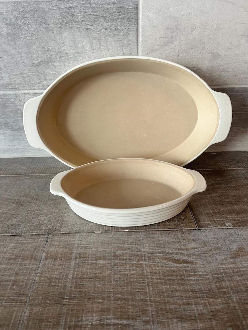 May include: Two oval ceramic baking dishes in off-white. The larger dish is behind a smaller one. Both have a light tan interior and white handles. The smaller dish has horizontal lines around the exterior.