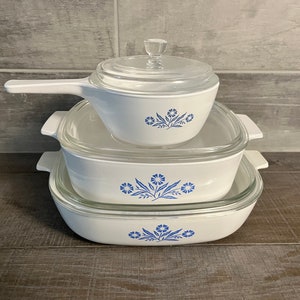 May include: A set of three white Pyrex baking dishes with blue floral designs. The dishes are stacked on top of each other, with the smallest dish on top. The dishes have glass lids.