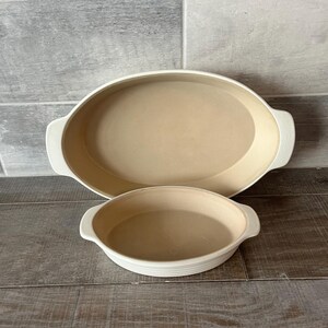 May include: Two off-white ceramic oval baking dishes. The larger dish is approximately 35 cm long, and the smaller dish is about 20 cm in diameter. Both have handles and a light tan interior.