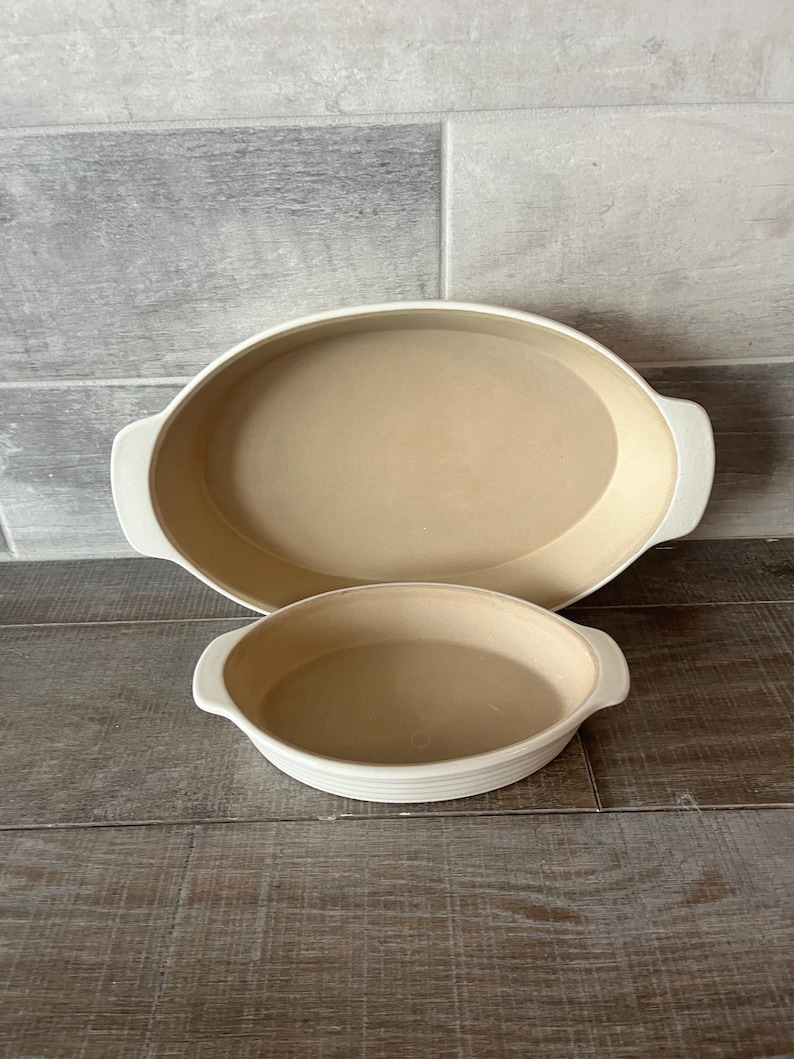May include: Two off-white ceramic oval baking dishes. The larger dish is approximately 35 cm long, and the smaller dish is about 20 cm in diameter. Both have handles and a light tan interior.