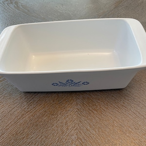 May include: White ceramic loaf pan with a blue floral design. The pan is empty and sitting on a wooden surface.
