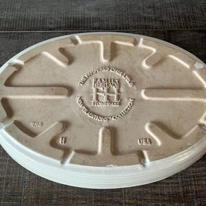May include: An off-white, oval stoneware baking dish. The base has embossed text: "THE PAMPERED CHEF LTD." and "FAMILY HERITAGE NEW TRADITIONS COLLECTION STONEWARE USA". The dish has a ridged edge.
