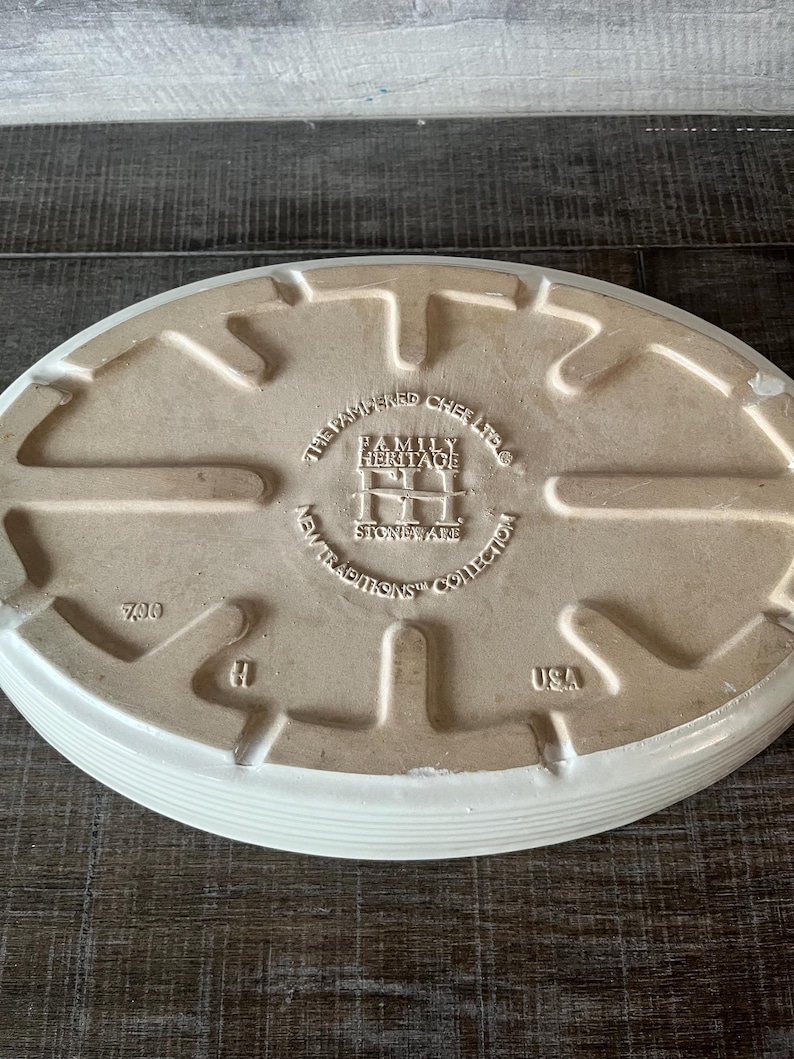 May include: An off-white, oval stoneware baking dish. The base has embossed text: "THE PAMPERED CHEF LTD." and "FAMILY HERITAGE NEW TRADITIONS COLLECTION STONEWARE USA". The dish has a ridged edge.
