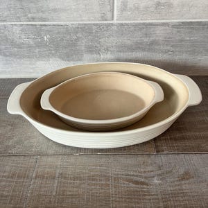 May include: Two off-white oval baking dishes, one nested inside the other. The larger dish has a ribbed exterior and handles. The smaller dish is smooth and fits snugly inside. Both are on a wooden surface.