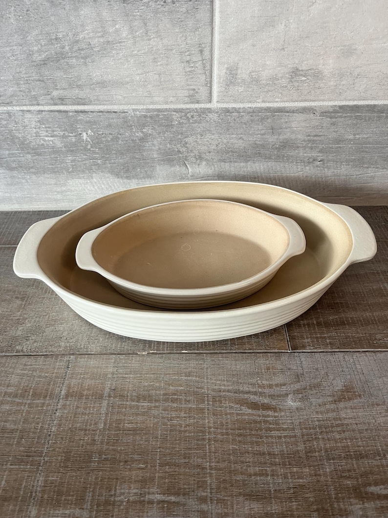 May include: Two off-white oval baking dishes, one nested inside the other. The larger dish has a ribbed exterior and handles. The smaller dish is smooth and fits snugly inside. Both are on a wooden surface.