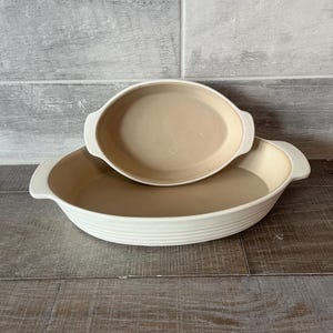 May include: Two oval, off-white ceramic baking dishes. The larger dish has a ribbed exterior and handles, while the smaller dish sits inside. Both have a tan interior. The background is a grey and white tiled wall and a wooden surface.