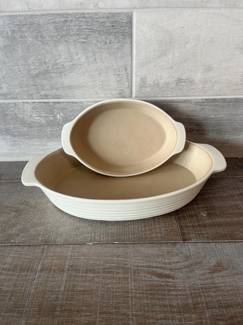 May include: Two oval, off-white ceramic baking dishes. The larger dish has a ribbed exterior and handles, while the smaller dish sits inside. Both have a tan interior. The background is a grey and white tiled wall and a wooden surface.