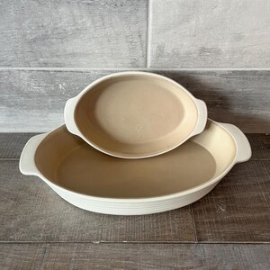 May include: Two oval beige ceramic baking dishes with white rims and handles. The larger dish is on the bottom, with a smaller dish nested on top. The dishes are on a wooden surface.