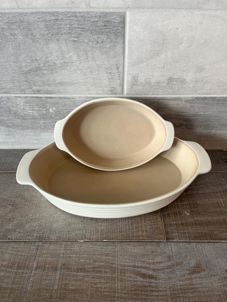 May include: Two oval beige ceramic baking dishes with white rims and handles. The larger dish is on the bottom, with a smaller dish nested on top. The dishes are on a wooden surface.