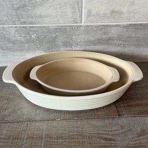 May include: Two ceramic baking dishes, one nested inside the other. The larger dish is off-white with a ribbed exterior and handles. The smaller dish is tan. Both are oval-shaped and sit on a wooden surface.
