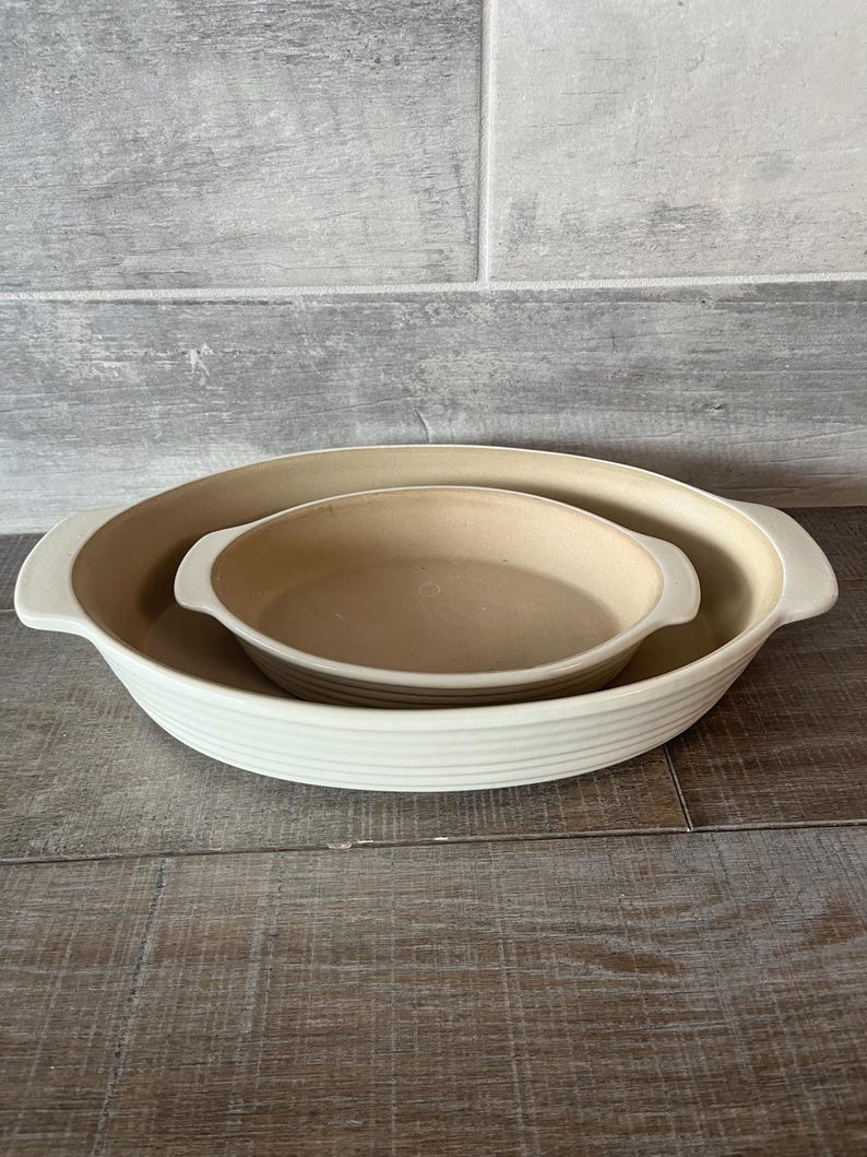 May include: Two ceramic baking dishes, one nested inside the other. The larger dish is off-white with a ribbed exterior and handles. The smaller dish is tan. Both are oval-shaped and sit on a wooden surface.