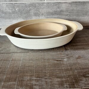 May include: Two ceramic baking dishes, one nested inside the other. The larger dish is white with a ribbed exterior and handles. The smaller dish is beige. The dishes are on a wooden surface.