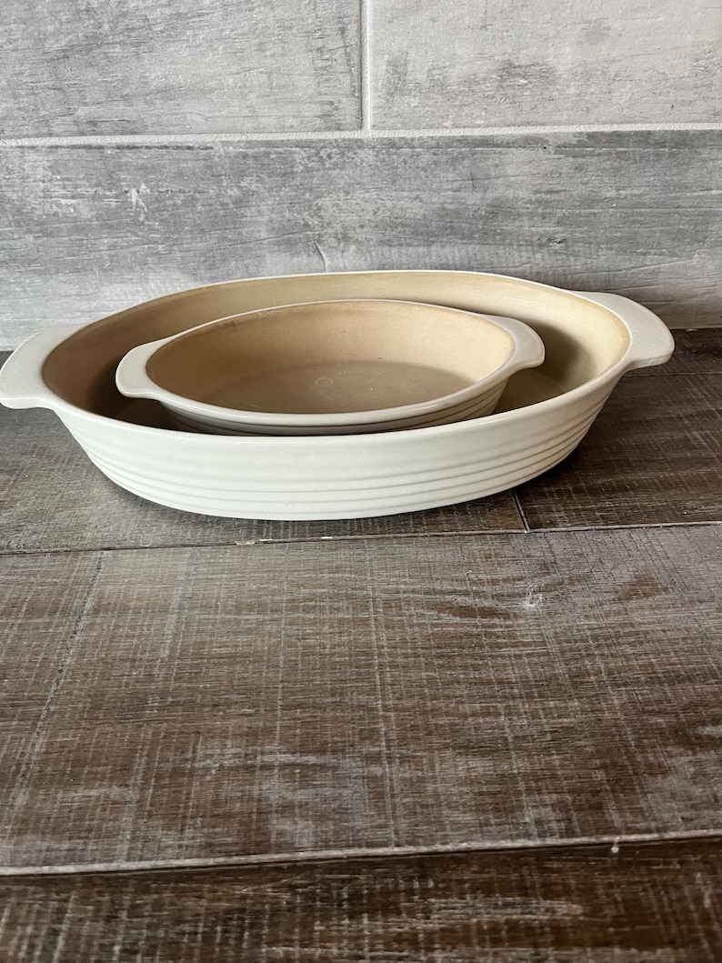 May include: Two ceramic baking dishes, one nested inside the other. The larger dish is white with a ribbed exterior and handles. The smaller dish is beige. The dishes are on a wooden surface.