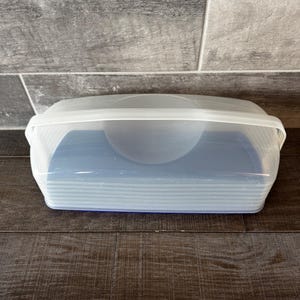 May include: A clear, rectangular food storage container with a white lid. The container has a blue interior and is sitting on a dark wood surface. The lid has a handle for easy opening and closing. The container is designed for storing food.