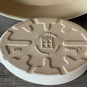 May include: An off-white, oval stoneware baking dish with a ridged edge. The base has embossed text: "The Pampered Chef Family Heritage Stoneware Collection" and "USA". A larger, round dish is partially visible in the background.