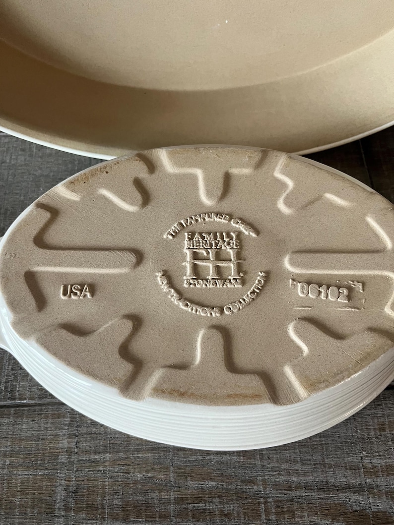 May include: An off-white, oval stoneware baking dish with a ridged edge. The base has embossed text: "The Pampered Chef Family Heritage Stoneware Collection" and "USA". A larger, round dish is partially visible in the background.