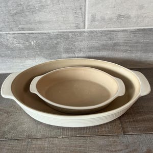 May include: Two beige oval baking dishes nested together. The larger dish has a ribbed exterior and white handles. The smaller dish is smooth with white handles. The dishes are on a wooden surface.