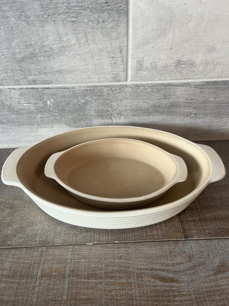 May include: Two beige oval baking dishes nested together. The larger dish has a ribbed exterior and white handles. The smaller dish is smooth with white handles. The dishes are on a wooden surface.