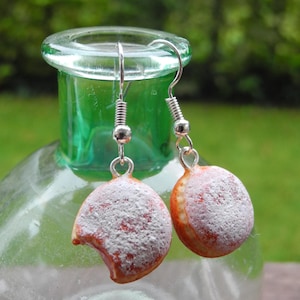 Könnte beinhalten: Ein Paar Ohrhänger in Form von Mini-Donuts. Die Ohrringe sind orange mit einem weißen Puderzuckereffekt und silberfarbenen Haken. Die Ohrringe werden vor einer grünen Glasflasche präsentiert.