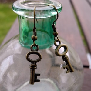 Könnte beinhalten: Ein Paar goldene Schlüssel-Ohrringe, die an einer durchsichtigen Glasflasche mit grünem Deckel hängen.