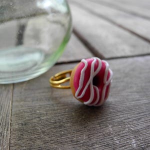 Könnte beinhalten: Ein Ring mit einem Miniatur-Donut-Anhänger, der eine rosa glasierte Oberfläche und weiße Zuckergussstreifen aufweist. Der Donut ist an einem dünnen, goldfarbenen Metallring befestigt. Das Design ahmt das Aussehen eines kleinen Gebäcks nach.